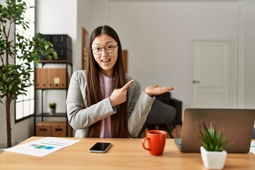 Sticker - Young chinese business worker wearing business style sitting on desk at office amazed and smiling to the camera while presenting with hand and pointing with finger.