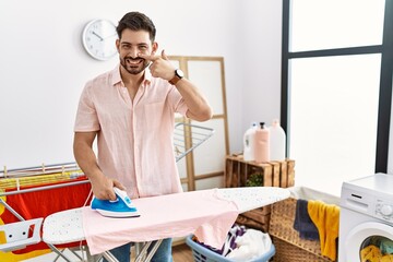 Poster - Young man with beard ironing clothes at home pointing with hand finger to face and nose, smiling cheerful. beauty concept