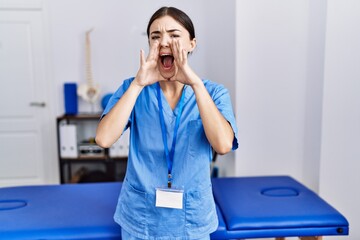 Poster - Young hispanic woman wearing physiotherapist uniform standing at clinic shouting angry out loud with hands over mouth