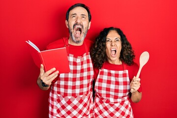 Middle age couple of hispanic woman and man wearing professional apron reading cooking recipe book angry and mad screaming frustrated and furious, shouting with anger looking up.