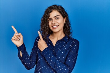 Poster - Young brunette woman with curly hair wearing casual clothes over blue background smiling and looking at the camera pointing with two hands and fingers to the side.