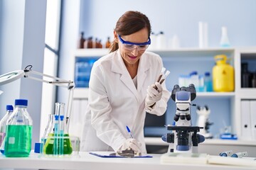 Poster - Middle age woman wearing scientist uniform writing on clipboard holding test tubes at laboratory