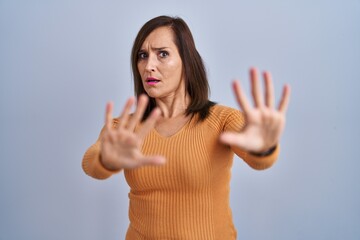 Poster - Middle age brunette woman standing wearing orange sweater afraid and terrified with fear expression stop gesture with hands, shouting in shock. panic concept.