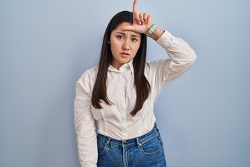 Canvas Print - Young latin woman standing over blue background making fun of people with fingers on forehead doing loser gesture mocking and insulting.