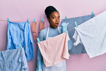 Poster - African american woman with braided hair washing clothes at clothesline surprised pointing with hand finger to the side, open mouth amazed expression.