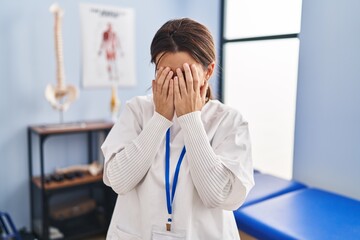 Canvas Print - Young brunette woman working at pain recovery clinic with sad expression covering face with hands while crying. depression concept.