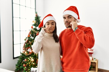 Poster - Young hispanic couple standing by christmas tree mouth and lips shut as zip with fingers. secret and silent, taboo talking