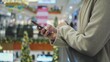 © rohawk - Young Caucasian Woman in Fashionable Clothes and Surgical Hygiene Mask Waiting in Shopping Mall Swiping Her Smartphone Looking for Best Offers on Christmas Season Sale Day