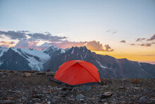 Scenic Alpine Landscape With Tent At Very High Altitude With View To Large Mountains In Orange Dawn Sky. Vivid Orange Tent With Awesome View To High Mountain Range Under Cloudy Sky In Sunset Colors.