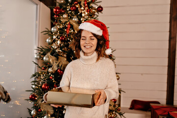 Wall Mural - Adorable cute girl with curls is posing at camera with happy smile and closed eyes holding a Christmas gift on background of Christmas tree