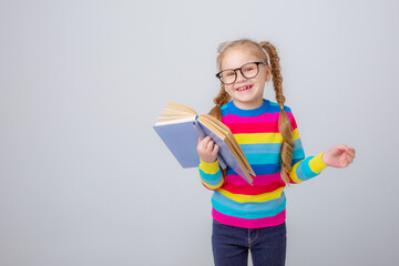 a cute little girl in a multicolored sweater and glasses holds a book on a white background , smiling