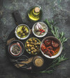 Various tasty Mediterranean food appetizer  in bowls : olives, capers, dried tomatoes , fresh rosemary herbs and olive oil served on wooden cutting board on dark rustic background. Top view