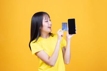 Wall Mural - Portrait Asian happy young girl smiling cheerful and showing plastic credit card while holding mobile phone isolated on yellow background