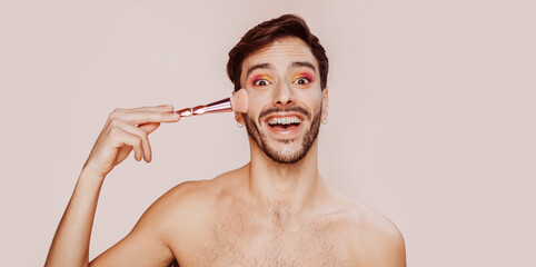 Portrait of cheerful smiling gay man with beard doing funny face, pink and yellow eyeshadow, no clothes, applying powder on face. Beauty, make-up and skin care concept. Copy space.