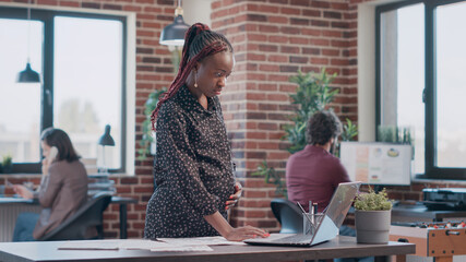 Wall Mural - African american woman expecting child and working on business project with documents. Pregnant employee analyzing files to design and plan marketing strategy for development at office.