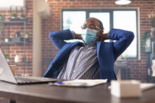 Employee Relaxing In Office After Finishing Business Project, Wearing Face Mask. Calm Entrepreneur Taking Break To Relax After Work And Getting Job Done. Man Sitting With Hands Over Head.