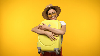 Wall Mural - Young traveler in sunglasses hugging suitcase isolated on yellow.