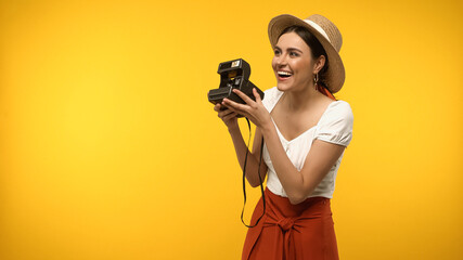 Wall Mural - Happy woman in straw hat holding vintage camera and looking away isolated on yellow.