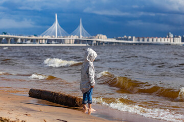 The child is standing on the seashore. Blue sky, waves, a bridge in the distance. A sunny summer day.