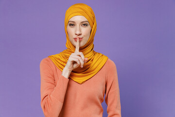 Wall Mural - Secret shocked amazed young arabian asian muslim woman in abaya hijab yellow clothes say hush be quiet with finger on lips shh gesture isolated on plain pastel light violet background studio portrait