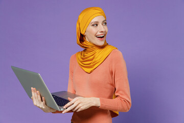 Wall Mural - Smiling vivid young arabian asian muslim woman in abaya hijab yellow clothes hold use work on laptop pc computer looking back behind isolated on plain pastel light violet background studio portrait.