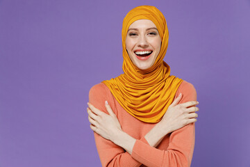 Wall Mural - Smiling vivid fun joyful overjoyed excited young arabian asian muslim woman in abaya hijab yellow clothes holding folded crossed hands isolated on plain pastel light violet background studio portrait.