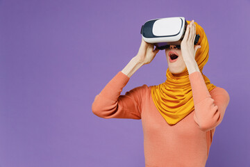 Wall Mural - Shocked amazed young arabian asian muslim woman in abaya hijab yellow clothes watching in vr headset pc gadget keeping mouth wide open isolated on plain pastel light violet background studio portrait.