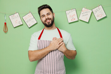 Poster - Young kind-hearted smiling happy male chef confectioner baker man 20s in striped apron put folded hands on heart isolated on plain pastel light green background studio portrait. Cooking food concept.