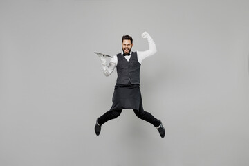 Wall Mural - Full body young male waiter butler man 20s in white shirt vest elegant uniform work at cafe jump high hold metal tray do winner gesture isolated on plain grey background . Restaurant employee concept.