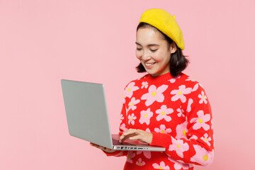 Wall Mural - Smiling happy woman of Asian ethnicity 20s in red knitted sweater beret hold use work on laptop pc computer isolated on plain pastel pink background studio portrait. People lifestyle fashion concept