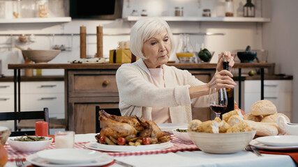 Wall Mural - elderly woman uncorking bottle of red wine near roasted turkey, grilled corn and buns.