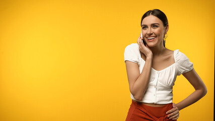 Wall Mural - Smiling woman in blouse holding hand on hip and talking on smartphone isolated on yellow.