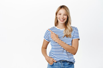 Wall Mural - Happy smiling blond woman pointing finger left, showing announcement or banner, announcing event, standing in tshirt over white background