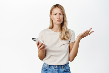 Wall Mural - Confused blond woman shrugging with mobile phone, cant understand smth on smartphone, standing puzzled over white background