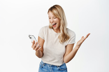 Wall Mural - Happy blond girl winning on mobile phone app, watching smartphone and reacting with joy and amazement to notification, standing over white background