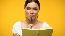 Sly Brunette Woman Holding Pen And Looking At Camera Near Notebook Isolated On Yellow.
