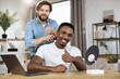 © sofiko14 - Portrait of stylish African man looking into the camera and showing thumb up while his Caucasian friend shaving his neck with an electric razor to create a stylish hairstyle. Multiracial gay couple
