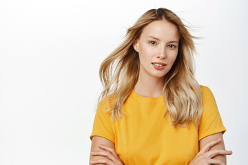 Wall Mural - Beauty and haircare. Attractive young woman with natural blond hair waving flying in air, looking stunning at camera, posing against white background in yellow t-shirt