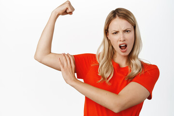 Wall Mural - Confident and sassy blond girl showing muscles, touching her arm, flexing biceps, strength and women power concept, standing over white background