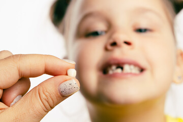 Wall Mural - Fingers holding torn out baby milk tooth with little blurred girl face looking at tooth and showing teeth behind on white background. First teeth changing. Going to dentist to do tooth treatment. 