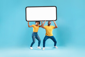 Wall Mural - black couple holding huge heavy smartphone with empty screen on blue background. mockup for ad, mobi