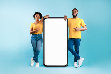 Wall Mural - great mobile app. african american couple pointing at giant smartphone with mockup, promoting app or