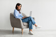 © Prostock-studio - Smiling young woman in casual wear working online, sitting in armchair and using laptop against white wall