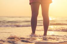 Close Up Woman Legs Walking On Tropical Sunset Beach With Smooth Wave And Bokeh Sun Light Wave Abstract Background. Travel Vacation And Freedom Feel Good Concept.