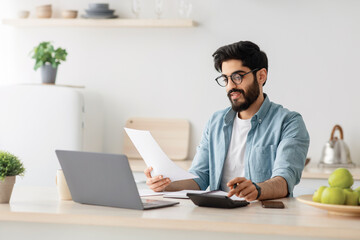 Home accounting, taxes. Happy young arab man in white minimalist kitchen interior, with computer and calculator