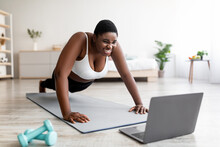 Plus Size Afro Woman Standing In Plank Pose Or Doing Push Ups Near Laptop, Working Out To Online Sports Tutorial At Home