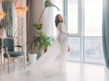 Young Beautiful Pregnant Stand Woman Near Window Hugs Stomach With Hands. White Long Silk Negligee Pregnancy Dress Peignoir, Gown Fabric Fly Wind, Flutter In Motion. Room Background. Happy Smile Face