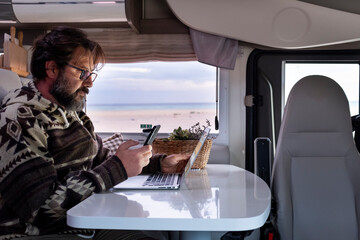 Wall Mural - Adult man working with laptop inside a camper van. Freedom remote worker lifestyle. Smart working with phone and roaming connection. Camping car interior and beach sunny  day outside the window 