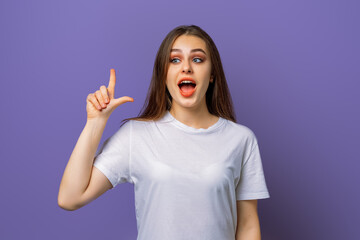 Wall Mural - Intrigued young woman with long hair pointing finger up, looking amazed aside, discussing promo above, standing over purple background