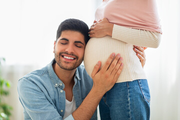 Wall Mural - Delighted eastern man embracing tummy of his pregnant wife, listening baby kicks, tenderly touching belly of his spouse